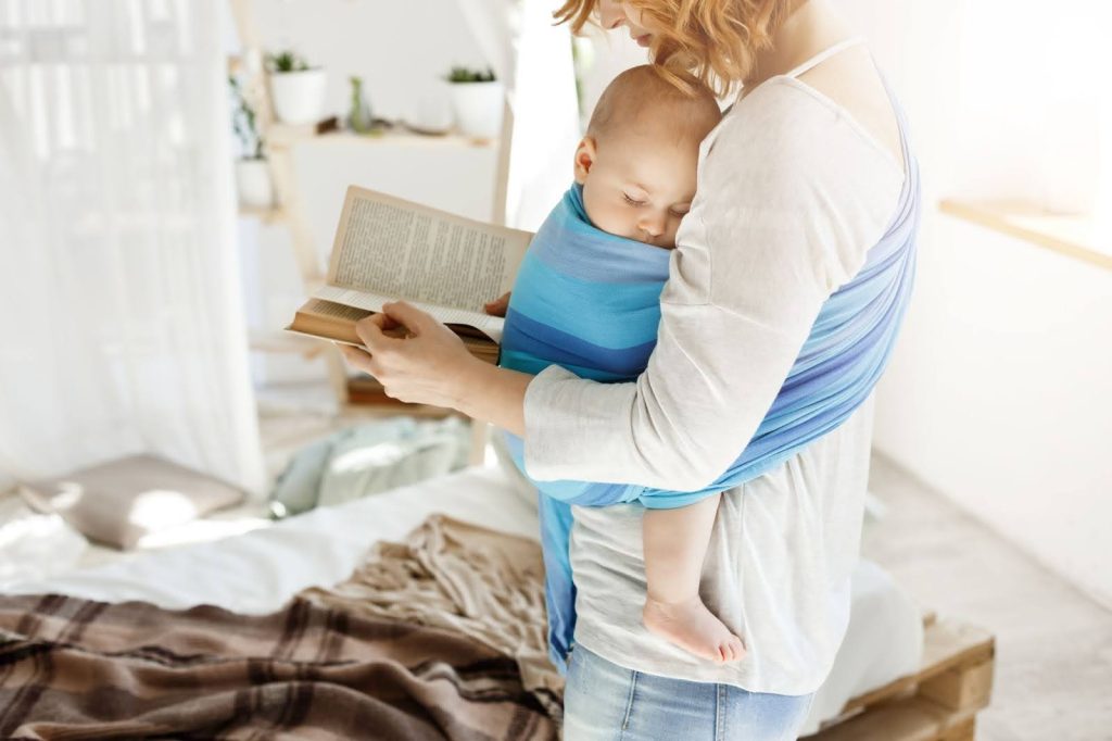 Ventajas y beneficios del porteo
Imagen de mujer realizando porteo con bebé utilizando un fular en tonos azules mientras lee